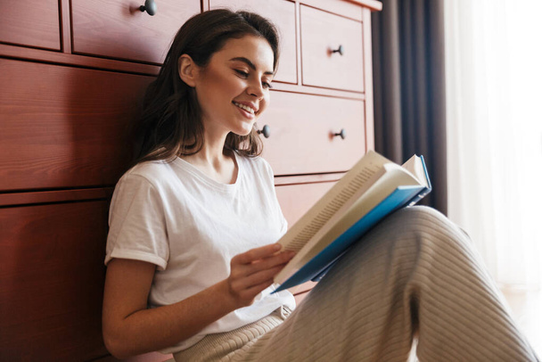 Attractive smiling young brunette woman reading a book while sitting on a floor and leaning on a cupboard - 写真・画像
