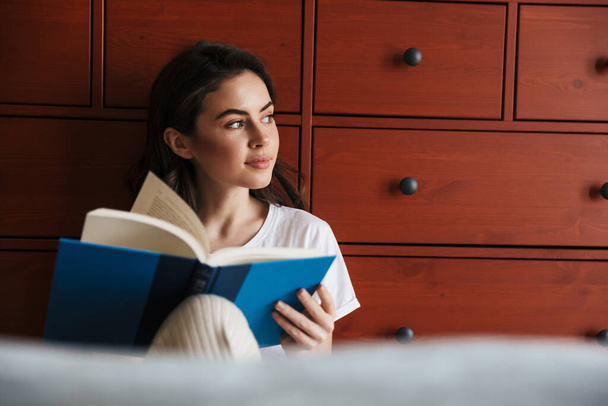 Attractive smiling young brunette woman reading a book while sitting on a floor and leaning on a cupboard - Photo, Image