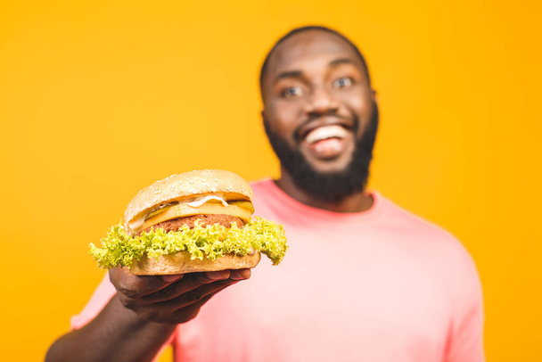 Young african american man eating hamburger isolated over yellow background. - 写真・画像