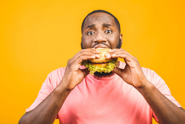 Young african american man eating hamburger isolated over yellow background. - Фото, изображение