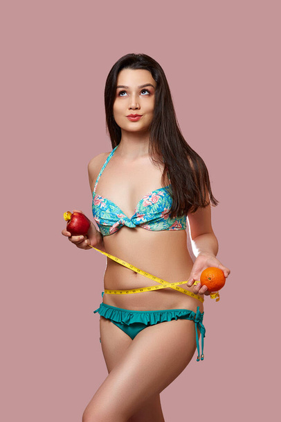 Μια νεαρή όμορφη μελαχρινή γυναίκα με μπλε μαγιό με ένα μήλο, ένα πορτοκάλι και μια μεζούρα στα χέρια της. σε ροζ απομονωμένο φόντο. - Φωτογραφία, εικόνα