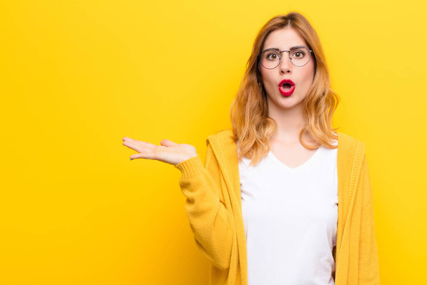 young pretty blonde woman looking surprised and shocked, with jaw dropped holding an object with an open hand on the side against yellow wall - Foto, Bild