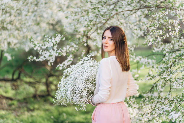春に開花する木の果樹園でポーズをとる帽子の素晴らしい若い女性。春の庭で香りを楽しむ美しい幸せな若い女性 - 写真・画像
