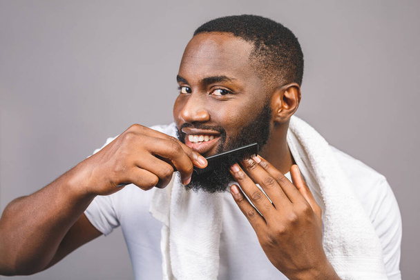 Egy fiatal, jóképű afro-amerikai fekete férfi portréja, aki a fürdőszobában fésüli a szakállát. Szürke háttérrel elszigetelve. - Fotó, kép
