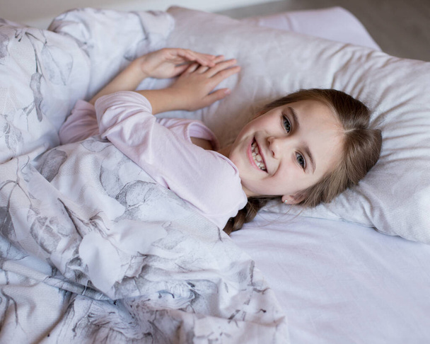 Cute little girl in bed woke up in the morning and smiles. Child lies on a pillow and looks at the camera. Pastime at home during quarantine. Home comfort. - Photo, Image