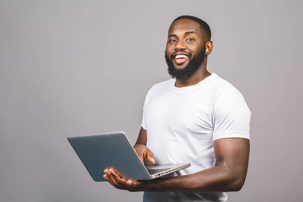 Fiatal mosolygós afro-amerikai férfi áll, és használja laptop elszigetelt szürke háttér. - Fotó, kép
