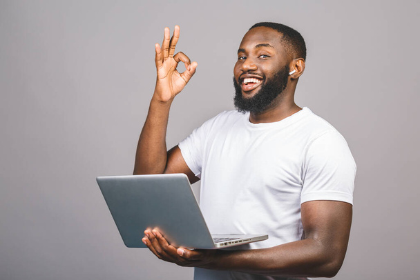 若い笑顔アフリカ系アメリカ人の男性が立って、灰色の背景に隔離されたラップトップコンピュータを使用しています。わかりましたサイン. - 写真・画像