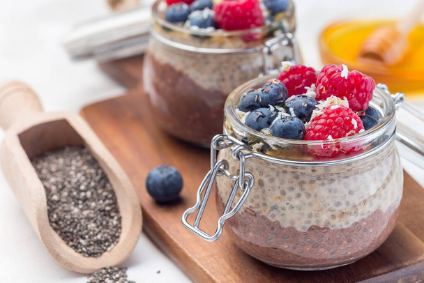 Layered chocolate and peanut butter chia seed pudding in a jar, garnished with raspberry, blueberry, honey and coconut flakes, horizontal - Foto, afbeelding