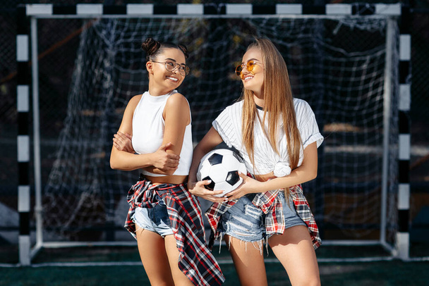 Dwie nowoczesne, młode hipsterki pozują ze szczęśliwymi twarzami przed bramką na stadionie piłkarskim.. - Zdjęcie, obraz