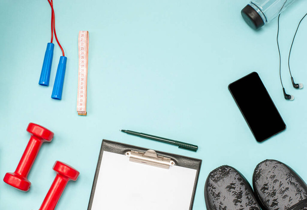 Flat lay of sports equipment for fitness on a blue background. The concept of a healthy lifestyle, sport and diet. Sports shoes, bottle of water, dumbbells, notepad, jump rope, phone, headphones. - Valokuva, kuva