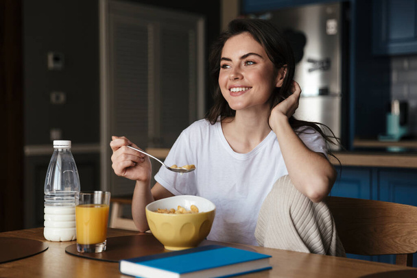 Attractive smiling young brunette woman having a healthy breakfast while sitting at the kitchen table - Foto, Bild