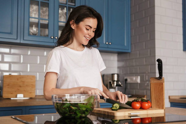 Beautiful young brunette woman cooking salad while standing at the kitchen counter - Foto, Imagen
