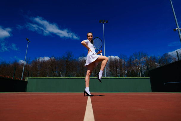 Σέξι τενίστρια με λευκό φόρεμα και τακούνια που κρατάει ρακέτα στο γήπεδο. Νεαρή γυναίκα παίζει τένις, σπορ. - Φωτογραφία, εικόνα