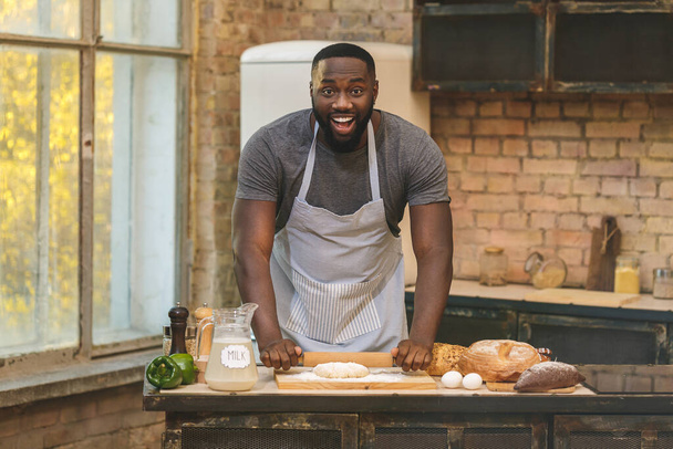Bakproces. Vrolijke donkere huid Afro-Amerikaanse man draagt schort, kneedt deeg met groot enthousiasme, tevreden te zijn met de lof van de chef-kok voor inspanning en hard werken in restaurant. - Foto, afbeelding