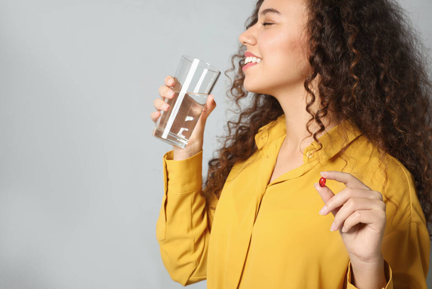 African-American woman with glass of water and vitamin capsule on light grey background - Фото, зображення
