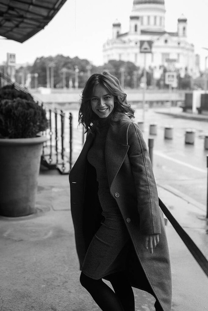 black and white portrait of young smiling happy pretty girl in coat, turtleneck posing outdoors. Street style portrait photo session of attractive female, urban photoshoot of beautiful elegant model - Фото, зображення