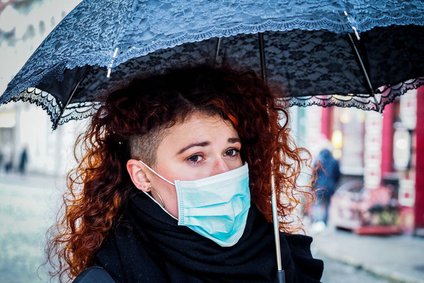 Beautiful young European woman in spring clothes on the street with a medical face mask on. Closeup of a 30-year-old female in a respirator to protect against infection with coronavirus - Covid19 - Photo, Image