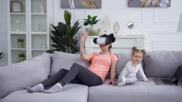 Close view of pretty confident 25-aged modern girl in virtual reality headset which sitting on the couch near her cute little child which looking cartoon on phone - Πλάνα, βίντεο