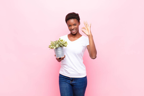 young pretty black womanfeeling happy, relaxed and satisfied, showing approval with okay gesture, smiling holding a plant - Fotoğraf, Görsel