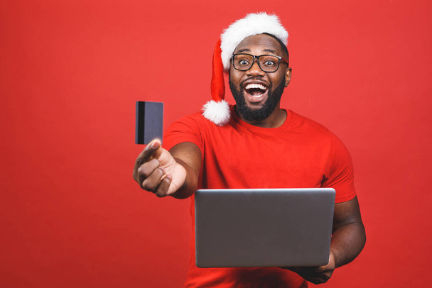 Porträt eines aufgeregten jungen afrikanisch-amerikanischen Mannes im Weihnachtsmannkostüm, der isoliert vor rotem Hintergrund steht, Laptop in der Hand hält und Plastikkreditkarte zeigt. - Foto, Bild