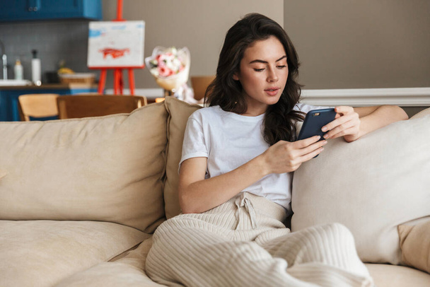 Beautiful young brunette woman relaxing on a couch at home,one using mobile phone - Фото, изображение