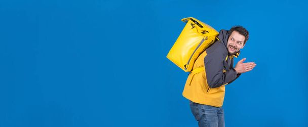 Emotional food delivery guy with grin on his face in yellow uniform and refrigerator bag on his back rubs his hands. Cunning courier wants to trick someone or do something to spite others - 写真・画像