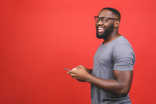 Portrait of handsome excited cheerful joyful delightful african american guy wearing casual sending and getting messages to his lover isolated against red background. Using phone.  - Фото, изображение