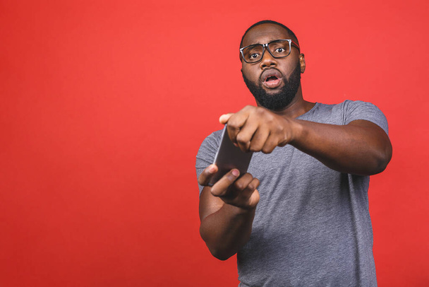 Portret van een vrolijke jonge zwarte Afro-Amerikaanse man gekleed in casual spelen games op mobiele telefoon geïsoleerd over rode achtergrond. - Foto, afbeelding