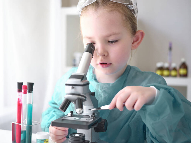 Χαριτωμένο κοριτσάκι που ψάχνει στο μικροσκόπιο στο γραφείο του στο σπίτι. Νεαρός επιστήμονας κάνει πειράματα στο σπίτι του εργαστηρίου.  - Φωτογραφία, εικόνα