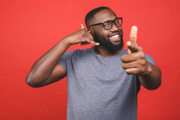 Mladý afroameričan v neformálním tričku stojí nad izolovaným červeným pozadím a usmívá se, mluví na telefonním gestu a ukazuje na vás. Zavolej mi.. - Fotografie, Obrázek