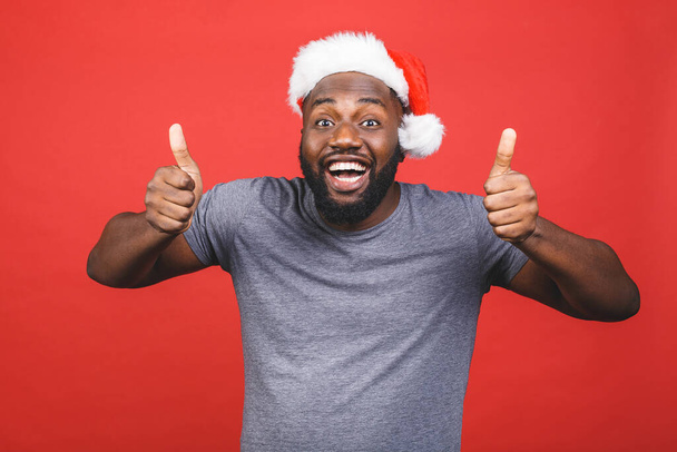 Nowy rok i czarny piątek wyprzedaży! Szczęśliwy uśmiechnięty czarny Afrykanin amerykański człowiek w świątecznym kapeluszu odizolowany na czerwonym tle. Kciuki w górę.  - Zdjęcie, obraz