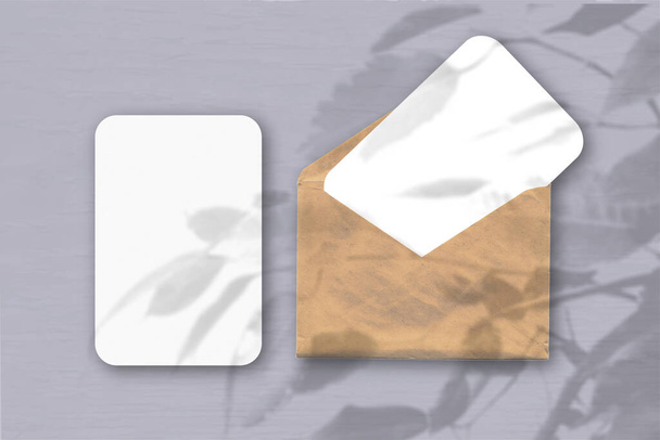 Um envelope com duas folhas de papel branco texturizado sobre um fundo de mesa cinza. Mockup com uma sobreposição de sombras de fábrica. A luz natural lança sombras de uma planta tropical
. - Foto, Imagem