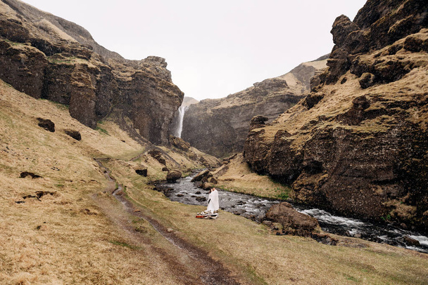 Destination Iceland wedding, near Kvernufoss waterfall. A wedding couple stands under a plaid near a mountain river. The groom hugs bride. They built an impromptu wedding table with decor and guitar - Foto, Imagen