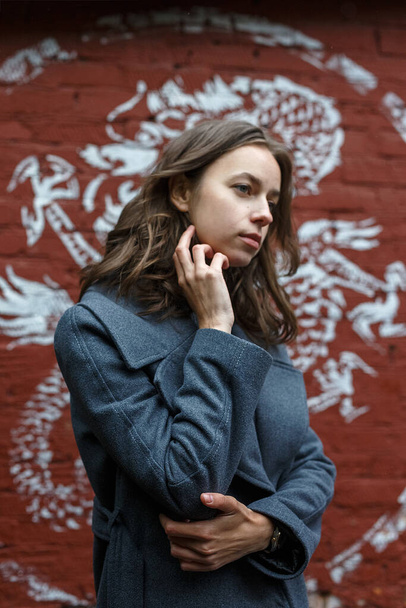 RUSSIA, MOSCOW, 10 JUN 2018: young thoughtful pretty girl in gray coat and turtleneck poses in front of red brick wall with graffiti. Street style portrait of female, urban photoshoot of elegant model - 写真・画像