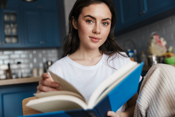 Attractive smiling young brunette woman reading a book while sitting on a kitchen - Фото, зображення