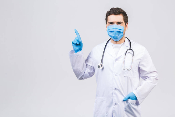 Doctor man in medical gown sterile face mask gloves isolated on white background. Epidemic pandemic coronavirus 2019-ncov sars covid-19 flu virus. Pointing finger aside up. - Foto, Imagen