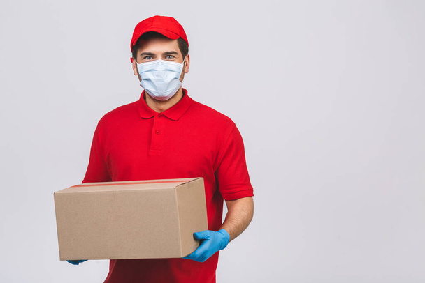 Dostawca pracownik w czerwonej czapce puste t-shirt jednolite rękawice maski twarzy trzymać puste pudełko kartonowe izolowane na białym tle. Koncepcja pandemii koronawirusa w służbie 2019-ncov. - Zdjęcie, obraz