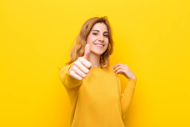 young pretty blonde woman feeling proud, carefree, confident and happy, smiling positively with thumbs up against yellow wall - Foto, Imagen