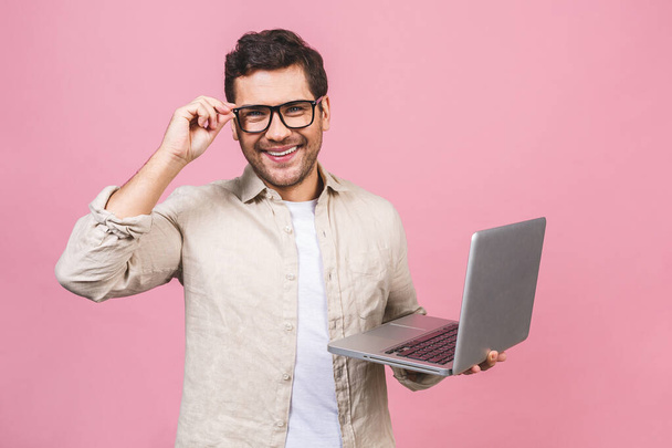 若いビジネスの男は、ピンクの背景に隔離された笑顔でコンピュータのラップトップを使用して作業眼鏡をかけて. - 写真・画像