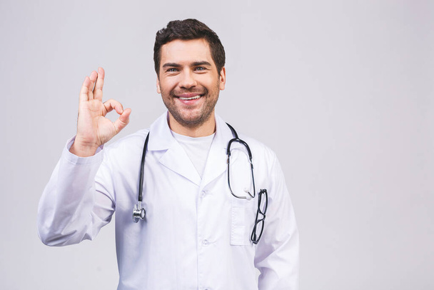 Portrait of handsome doctor in white coat, showing Ok sign, looking at camera and smiling, isolated on white background. - Foto, imagen
