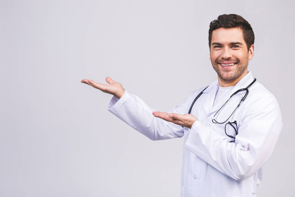 Jeune homme souriant médecin, professionnel de la santé tenant quelque chose dans la main vide isolé sur fond blanc. Pointant du doigt
.  - Photo, image