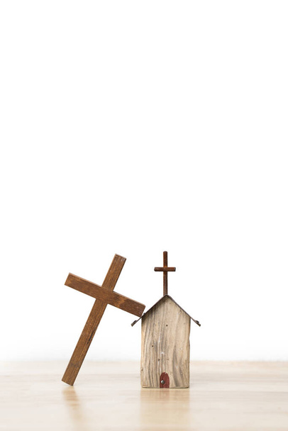 Деревянный крест, опирающийся на деревянный макет домика на деревянной доске и белом фоне
 - Фото, изображение