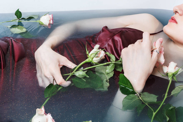 Above shot of young adult female in red dress lying in bathtub with blue water and roses - Foto, afbeelding
