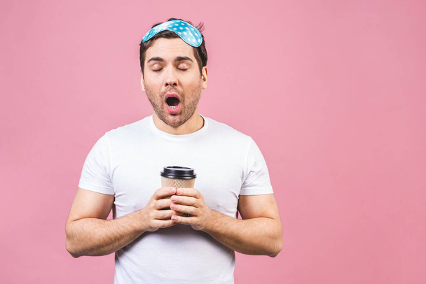 Portrait of nice calm peaceful sleepy young man with blue sleeping mask and cup of coffee or tea. Isolated over pink background. - Foto, imagen