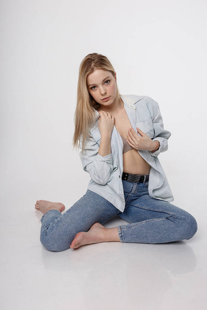 portrait of sexy caucasian woman with long hair posing in blue shirt and jeans on white studio background. model tests of pretty girl in basic clothes. attractive female sitting on floor on her knees - Фото, зображення