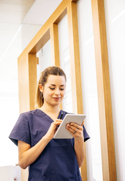 Низкий угол работы молодой женщины в униформе, улыбающейся и просматривающей данные на смартфоне во время работы в современной клинике - Фото, изображение