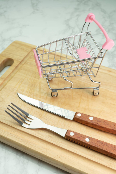 Carro de compras sobre tabla de cortar de madera con tenedor y cuchillo
 - Foto, Imagen