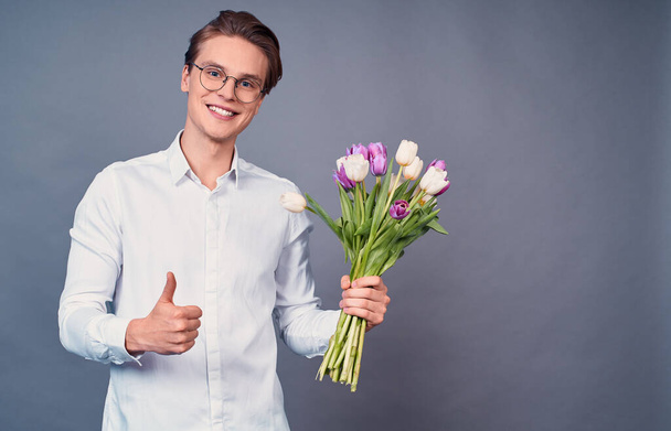 Young handsome man, in a white shirt and glasses, holds white and pink tulips, smiles happily, thumb up with fingers, excellent sign. Copy space. Delicate tulips for her. - Foto, Imagem
