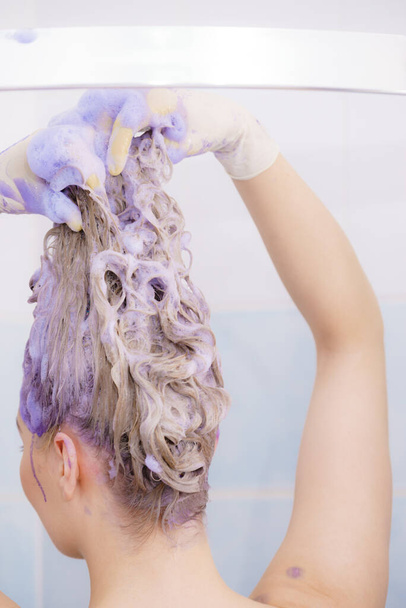 Woman applying coloring shampoo on her hair. Female having purple washing product. Toning blonde color at home. - 写真・画像