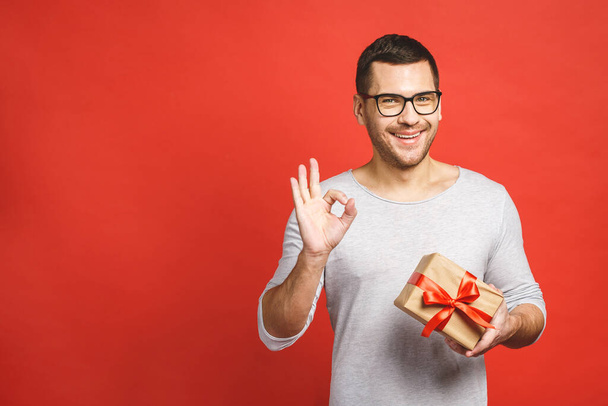 Happy holiday, my congredulations! Portrait of an attractive casual man giving present box and looking at camera isolated over red background. - Foto, afbeelding